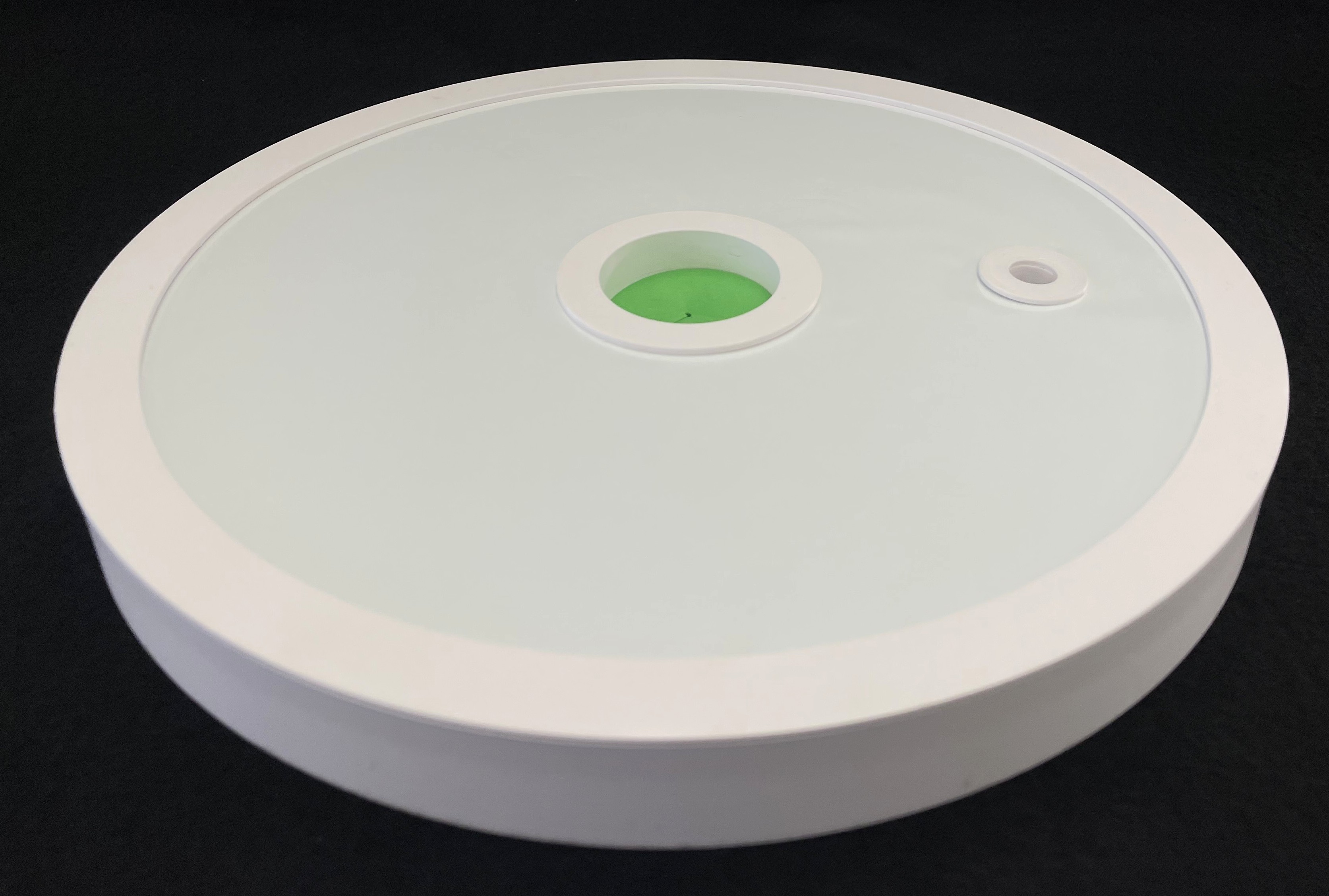 A whitle plastic lid with a green foam circle submerged in the center and a small hole to the right of the green circle.