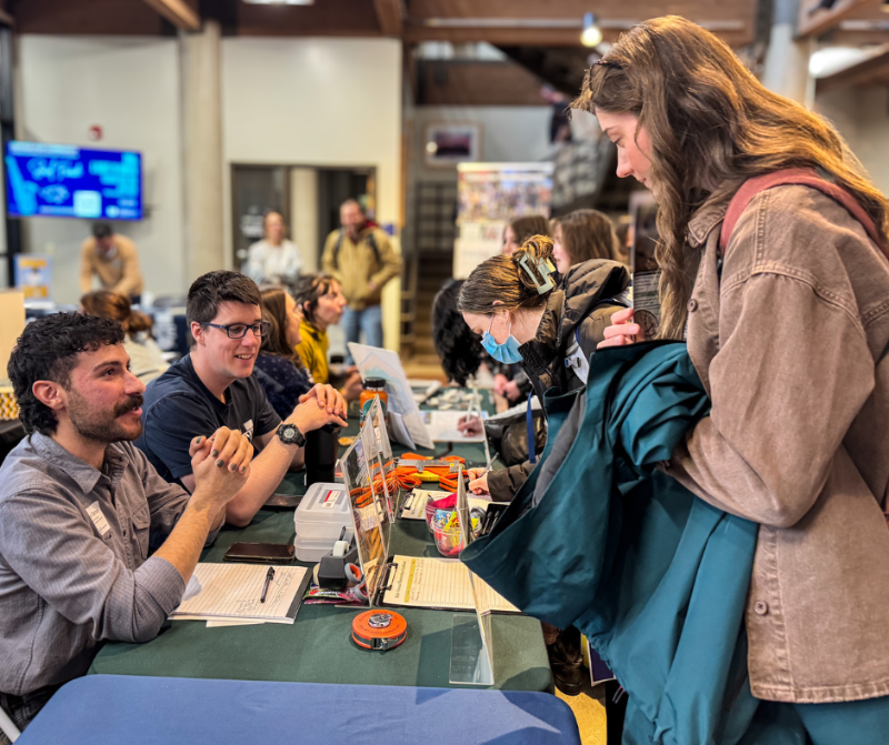 Two men sit at a table at a job fair talking to a woman who is standing.